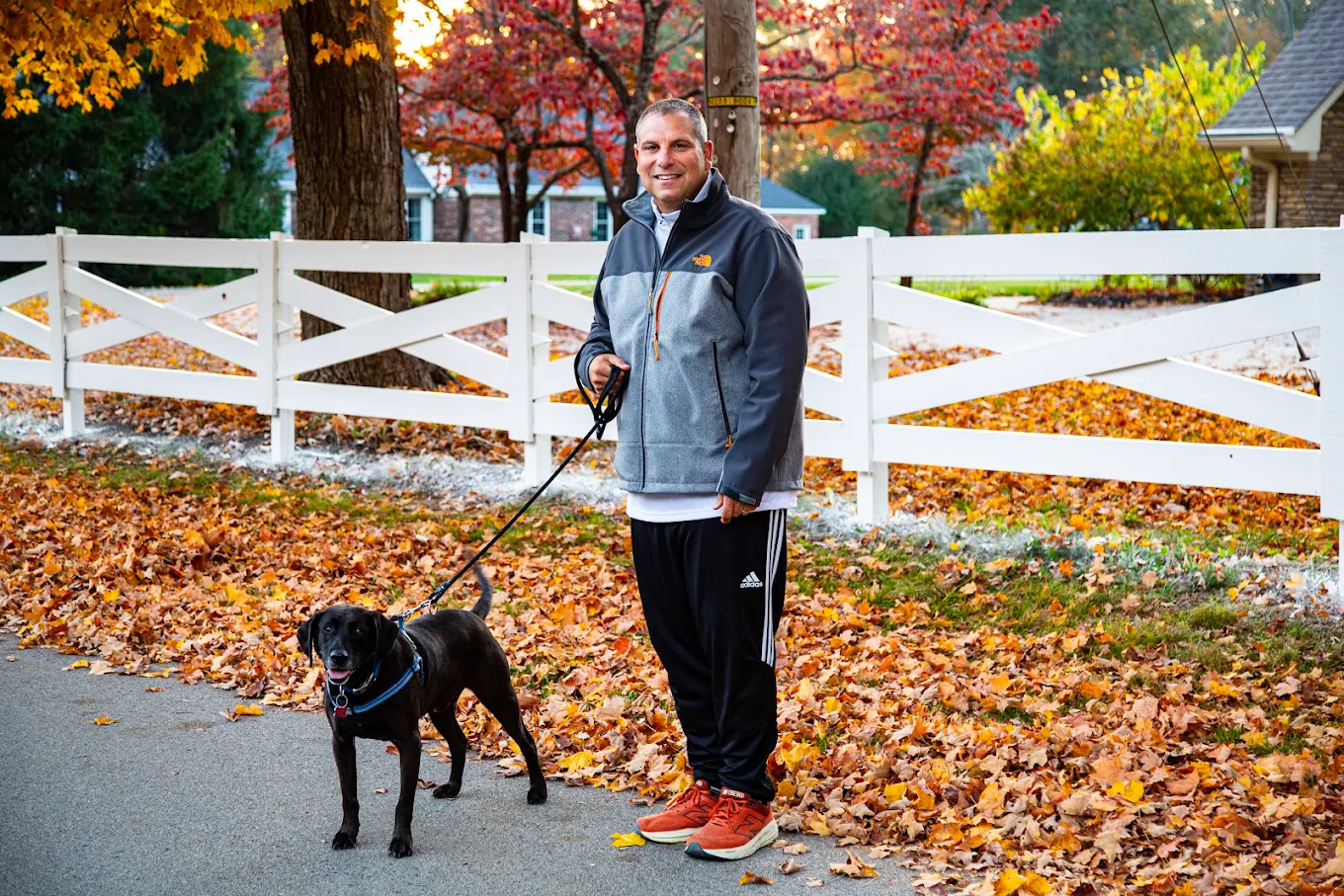 A man walking his black labrador on a leash along a leaf-strewn street in autumn
